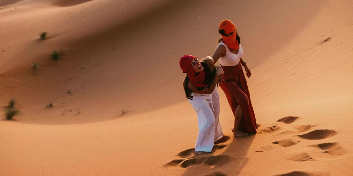 Two Contiki travellers climbing the golden sand dunes of the Sahara Desert in Morocco, dressed in traditional desert scarves during a sunset adventure on the Morocco Uncovered Tour