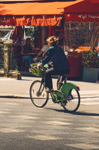 Woman cycling on a bike past a classic red-awninged French café terrace on a sunny Paris street - Contiki Europe Tour