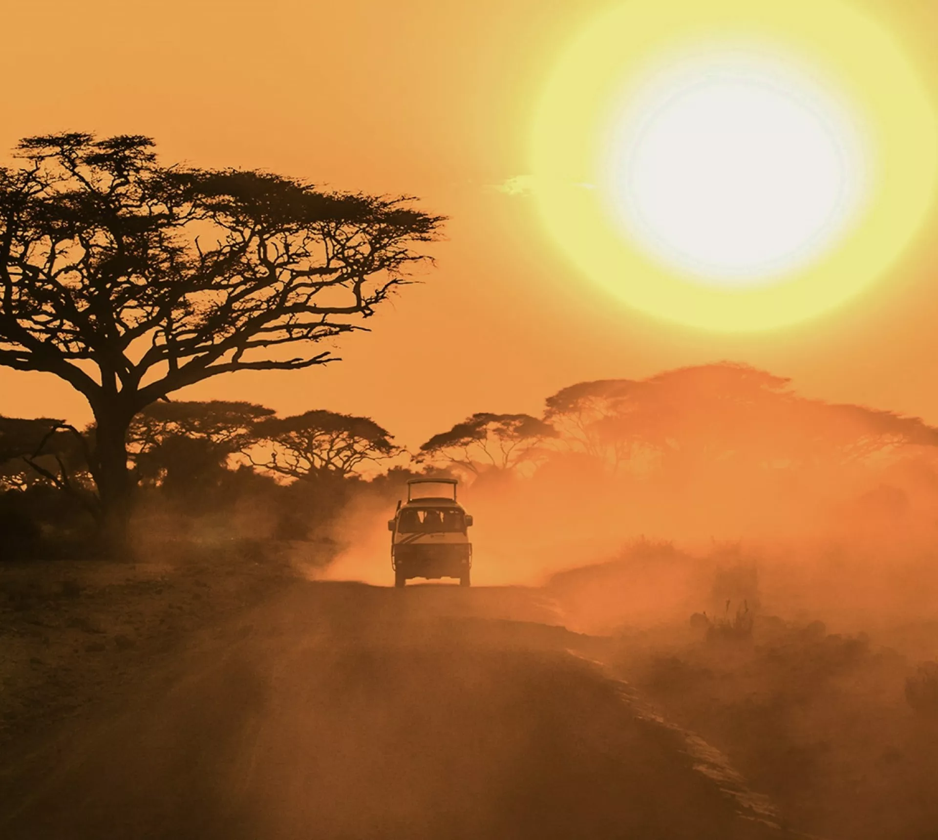 A safari vehicle drives along a dusty road beneath acacia trees as the sun sets over the East African savannah.