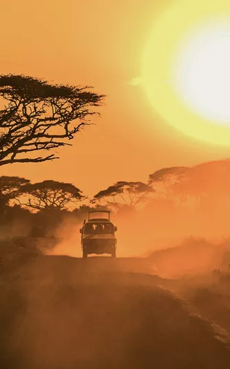 A safari vehicle drives along a dusty road beneath acacia trees as the sun sets over the East African savannah.