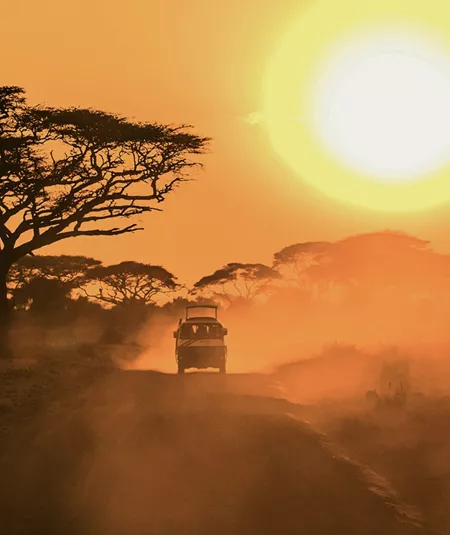 A safari vehicle drives along a dusty road beneath acacia trees as the sun sets over the East African savannah.