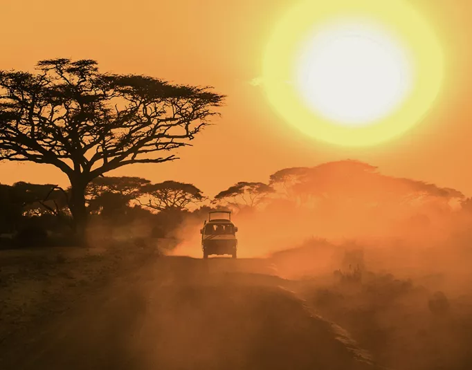 A safari vehicle drives along a dusty road beneath acacia trees as the sun sets over the East African savannah.