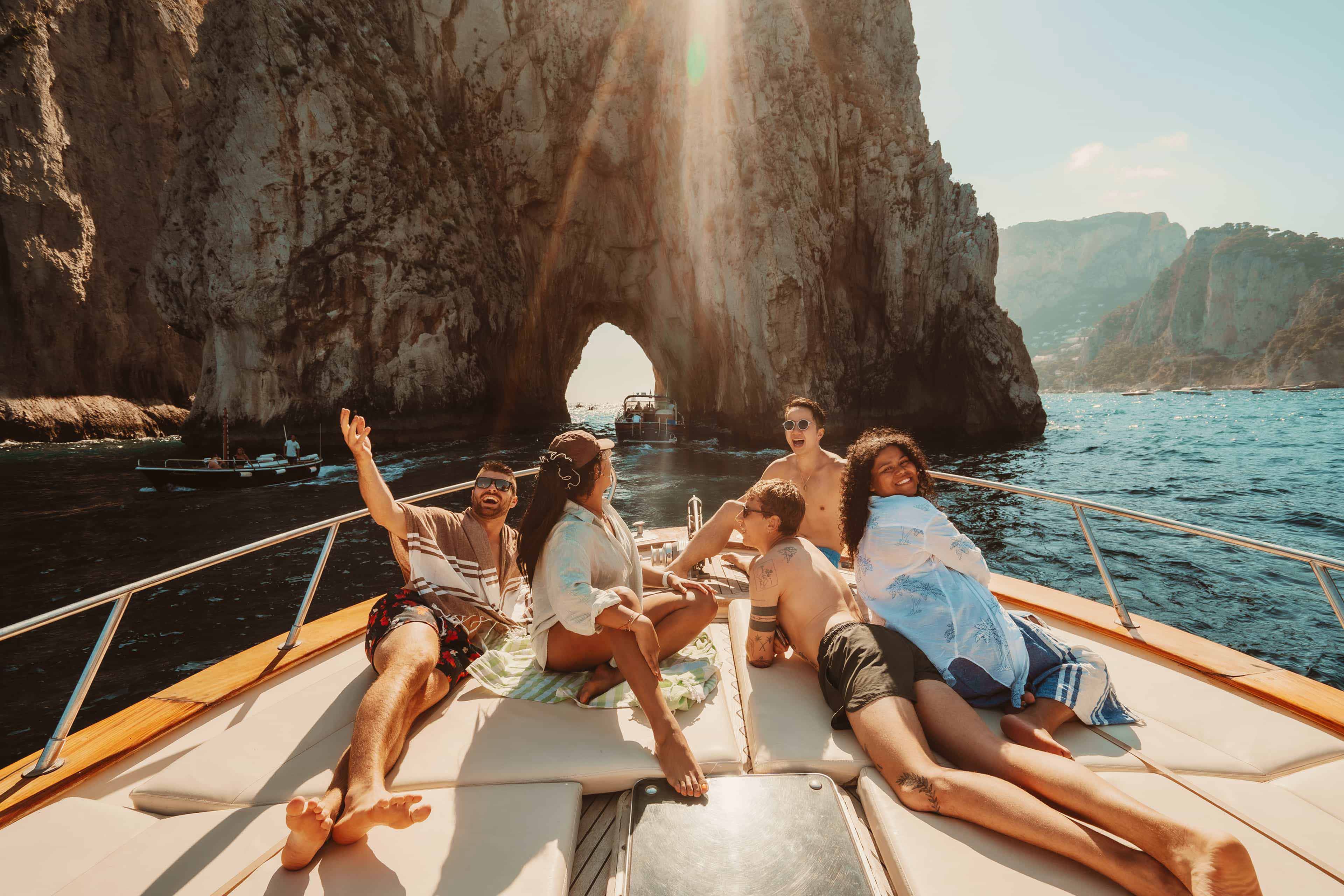 Friends laugh and relax on the bow of a boat near the Faraglioni sea arch off Capri, Italy.