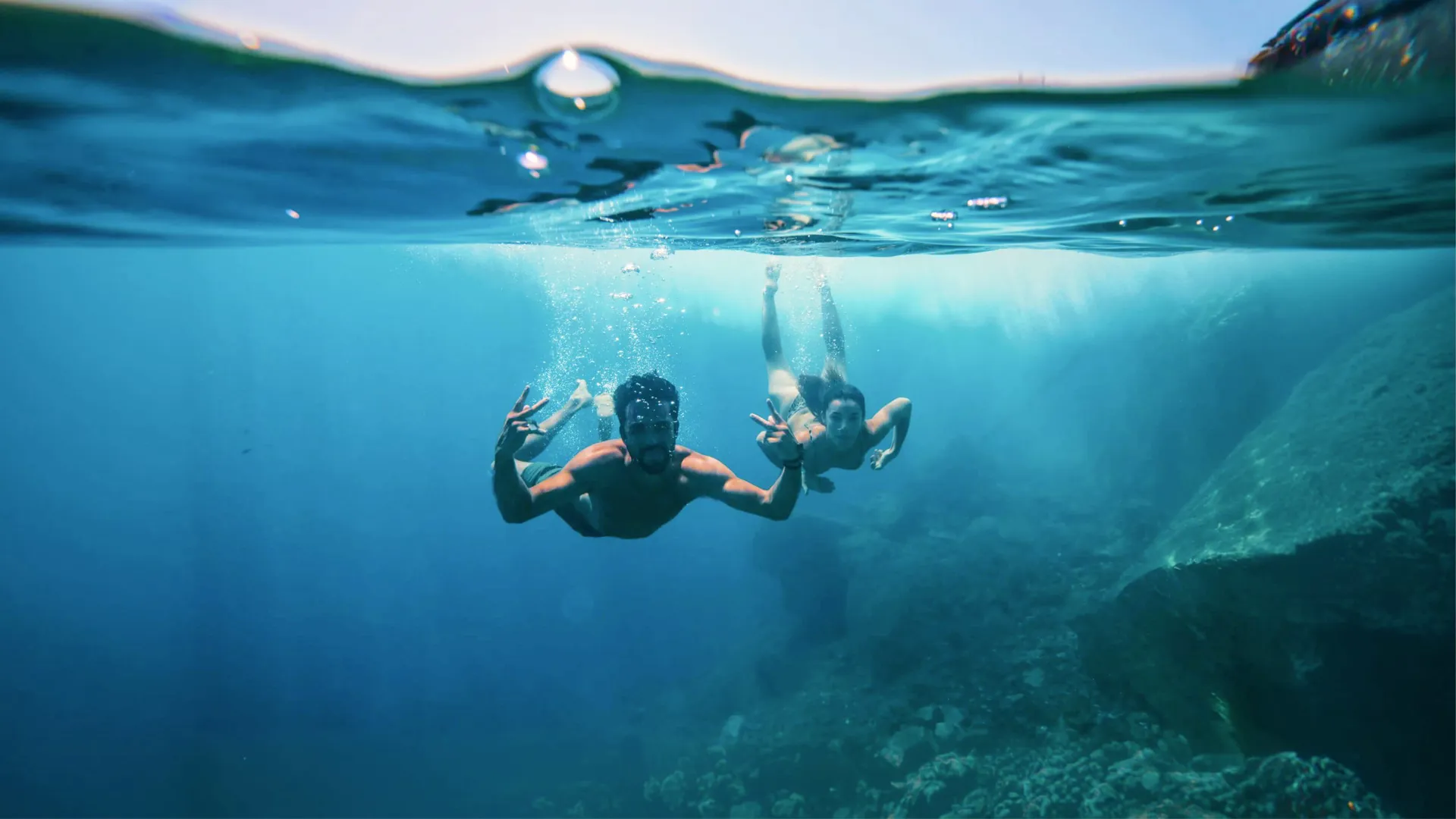 Two young travellers freediving underwater making peace signs in the clear blue waters of the Greek Islands - Contiki Athens and Santorini island hopper Tour