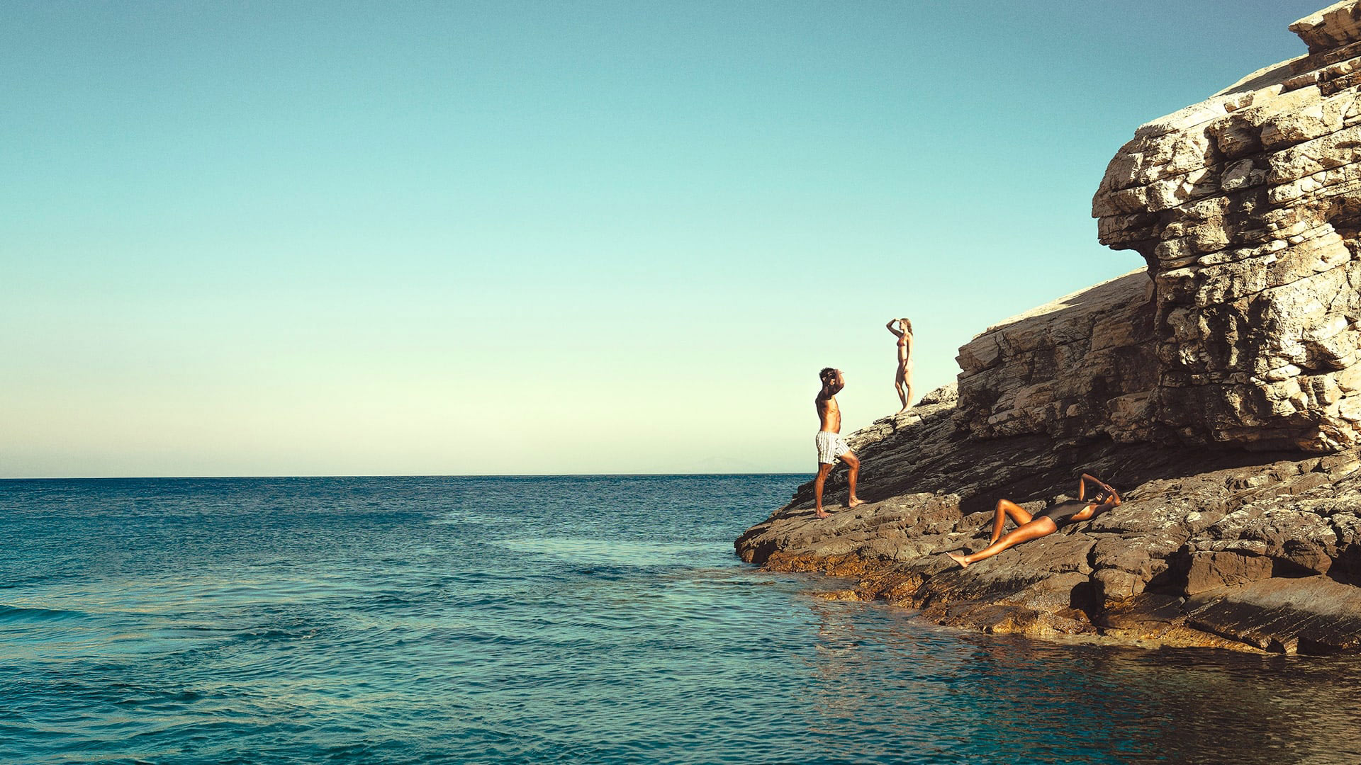 Travellers relax and take in the view from sun-warmed rocky cliffs above the sea in the Greek Islands.