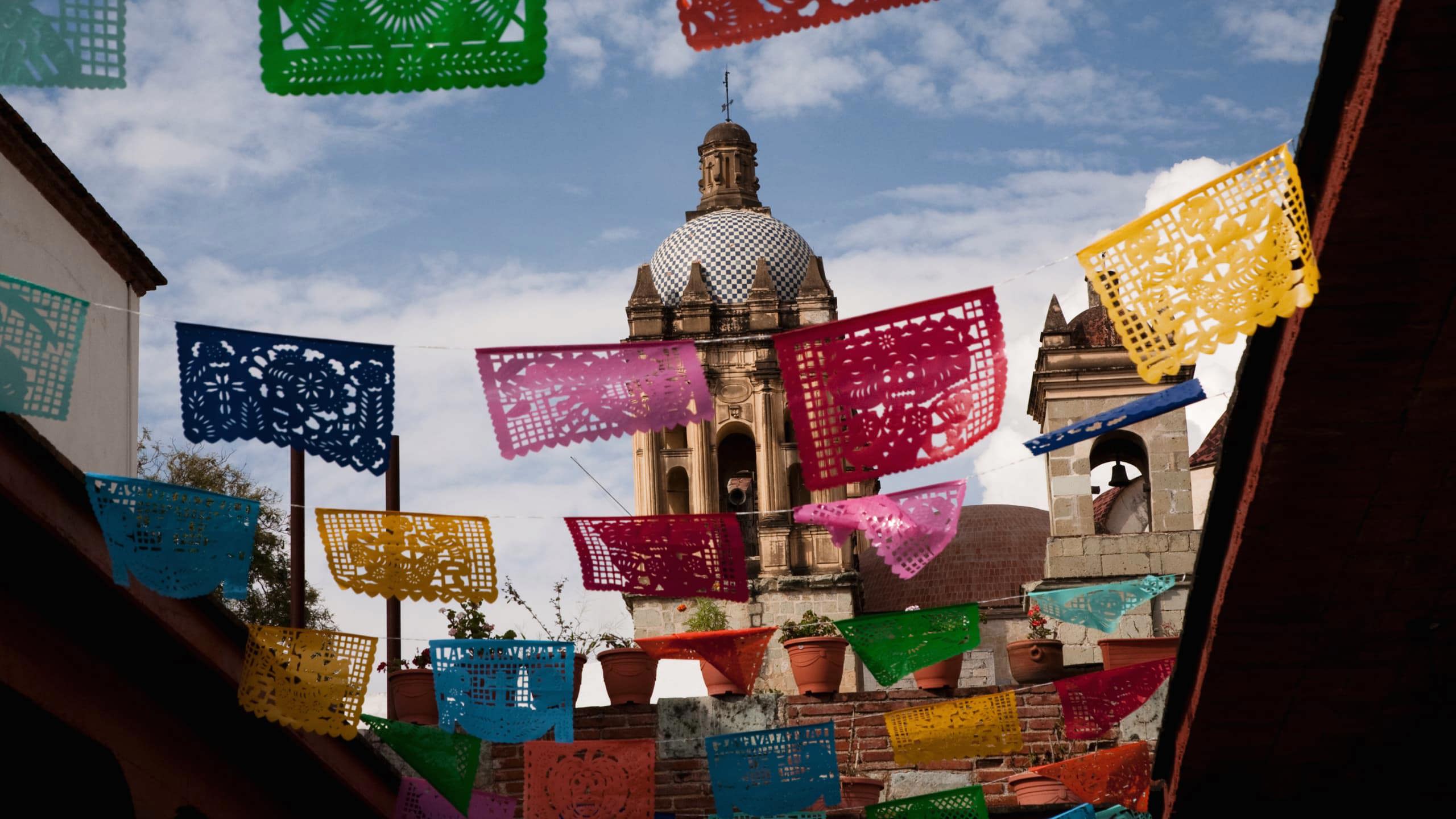 Colourful papel picado banners hanging above a colonial Mexican street with a baroque church dome and bell tower in Oaxaca Mexico - Contiki Mexico Tour