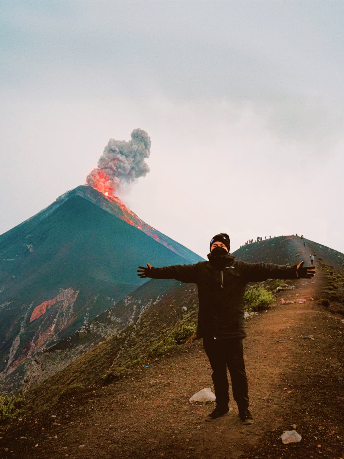 Traveller with arms outstretched on a ridge overlooking an erupting Fuego volcano in Guatemala.