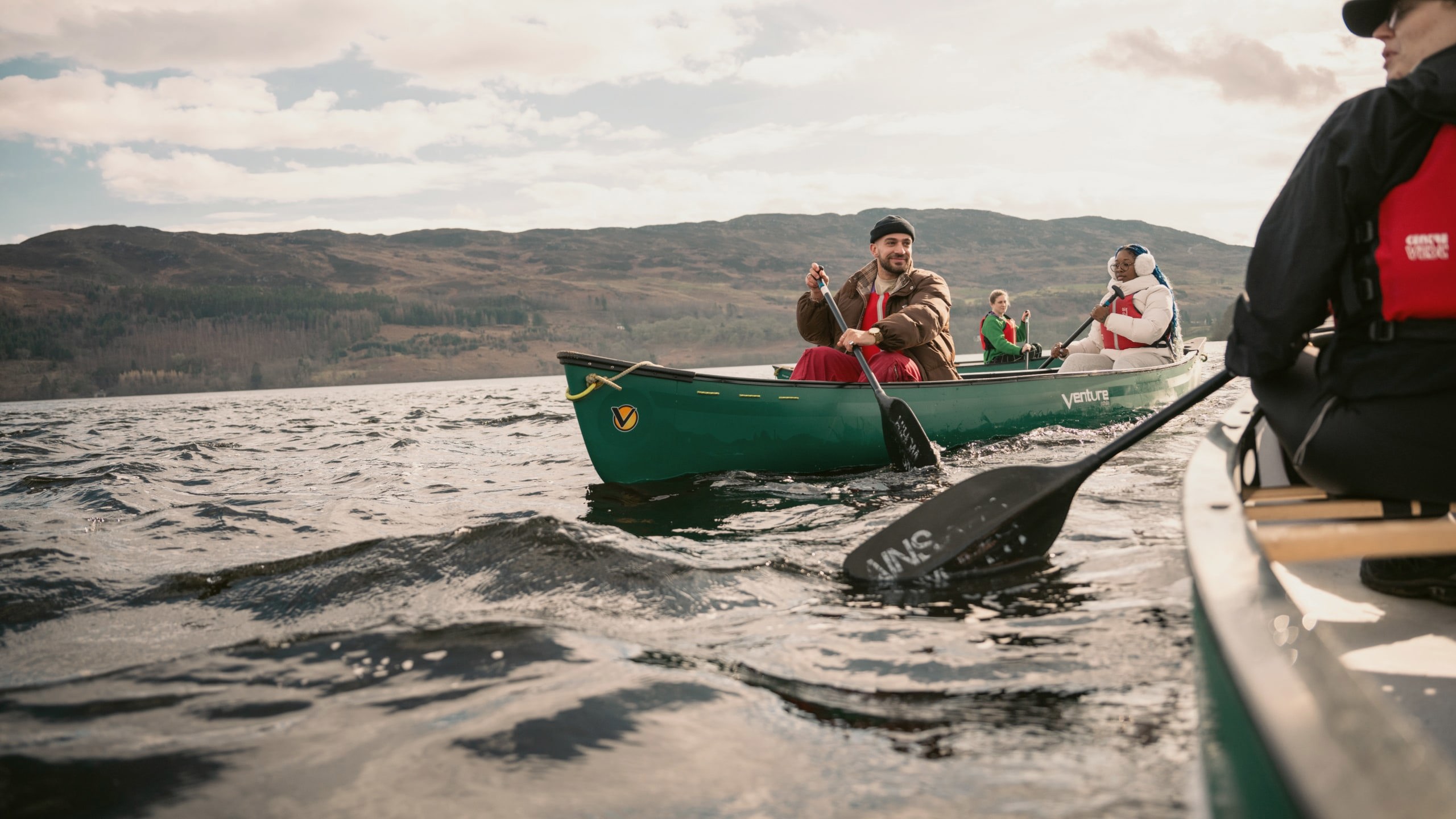 Contiki Travellers explore the Scottish Highlands by canoe, paddling across a tranquil loch with rolling hills in the background - Best of England & Scotland Summer Tour