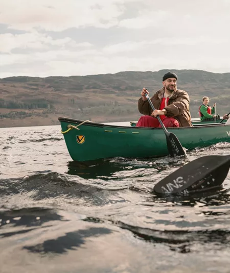 Contiki Travellers explore the Scottish Highlands by canoe, paddling across a tranquil loch with rolling hills in the background - Best of England & Scotland Summer Tour