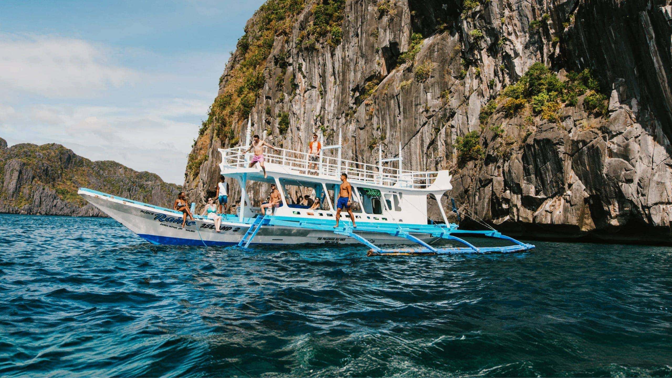 Group of young travellers jumping off a traditional Filipino bangka boat into the sea surrounded by dramatic limestone cliffs in El Nido Palawan Philippines - Contiki Philippines island hopping Tour