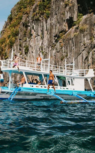 Group of young travellers jumping off a traditional Filipino bangka boat into the sea surrounded by dramatic limestone cliffs in El Nido Palawan Philippines - Contiki Philippines island hopping Tour
