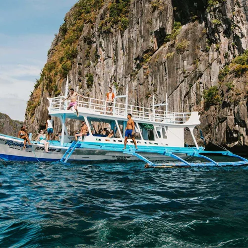 Group of young travellers jumping off a traditional Filipino bangka boat into the sea surrounded by dramatic limestone cliffs in El Nido Palawan Philippines - Contiki Philippines island hopping Tour