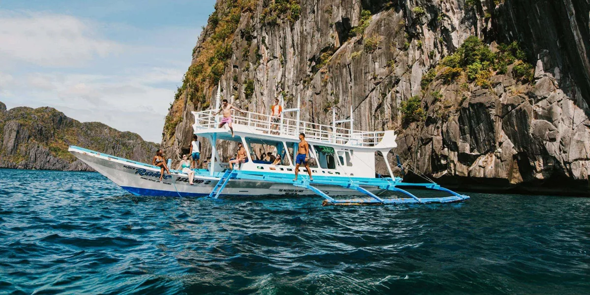 Group of young travellers jumping off a traditional Filipino bangka boat into the sea surrounded by dramatic limestone cliffs in El Nido Palawan Philippines - Contiki Ultimate Philippines Island Hopping Tour