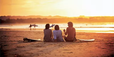 Contiki travellers relaxing on the sand with surfboards watching surfers walk along the shoreline at a stunning golden sunset in Byron Bay Australia
