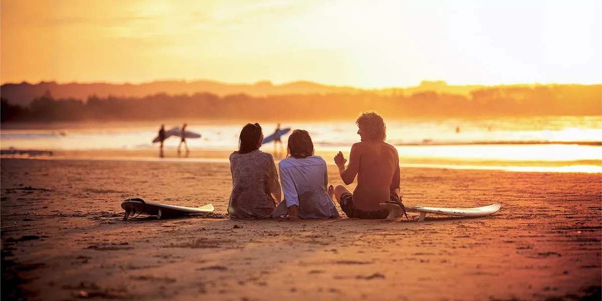 Contiki travellers relaxing on the sand with surfboards watching surfers walk along the shoreline at a stunning golden sunset in Byron Bay Australia