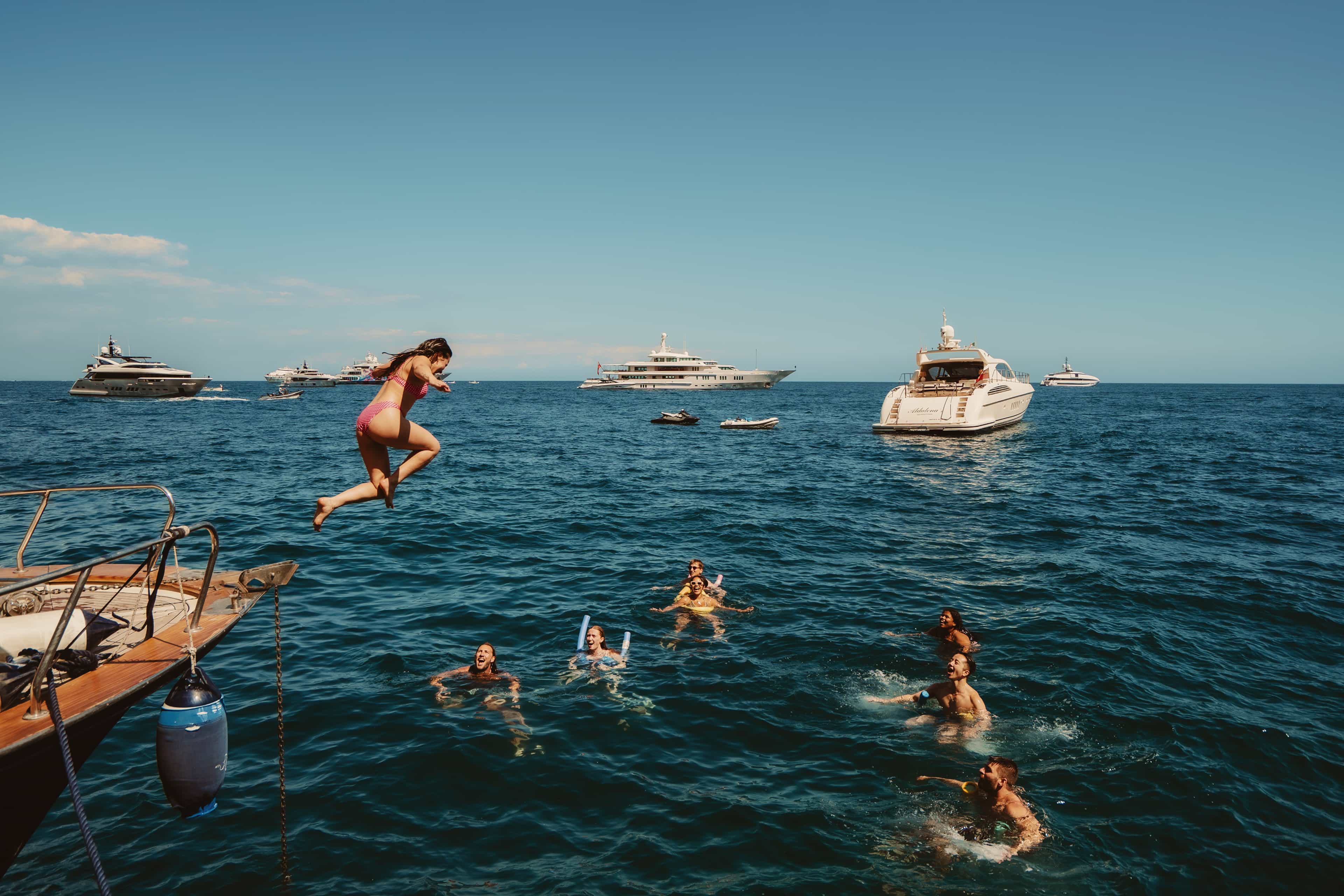 A traveller jumps from a boat into the Mediterranean Sea to join friends swimming, with yachts dotting the water behind them.