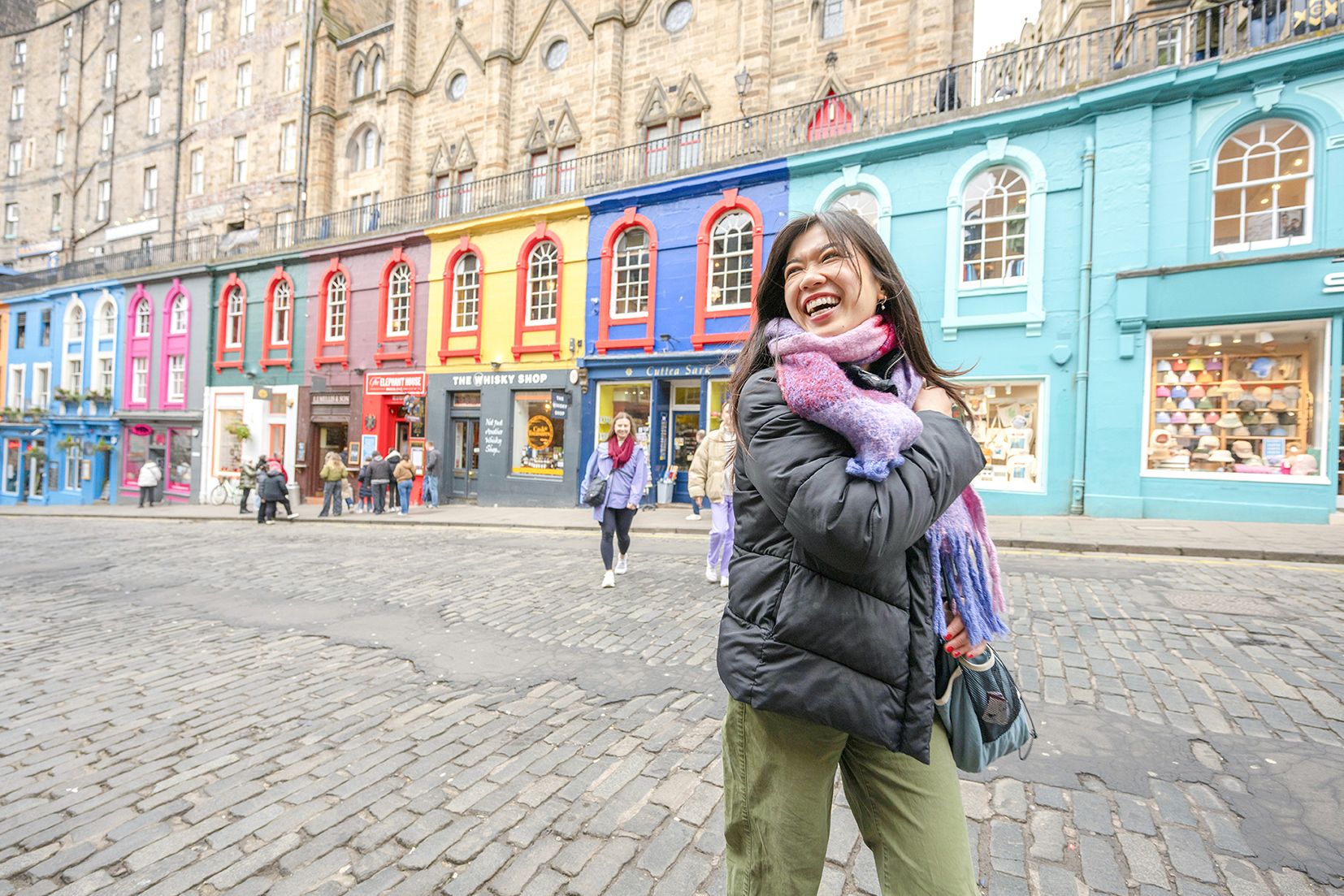 A traveller smiling standing in front of a colourful row of shops in Edinburgh, Scotland