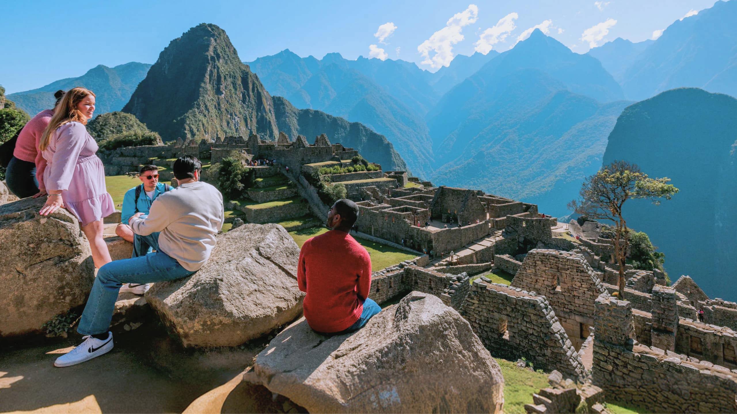 Group of young Contiki travellers sitting on rocks overlooking the ancient Inca ruins of Machu Picchu with Huayna Picchu mountain in the background Peru - Contiki South America Lima to Rio Tour