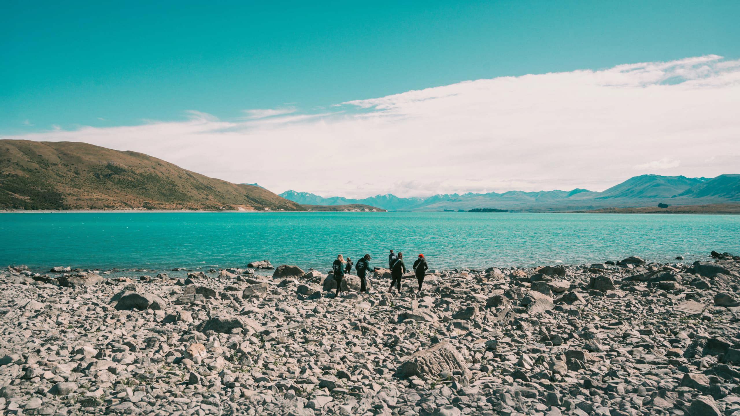 Group of travellers walking along the rocky shoreline of glacial Lake Pukaki with the vivid turquoise water and snow-capped Southern Alps in the background New Zealand - Contiki Best of NZ Tour
