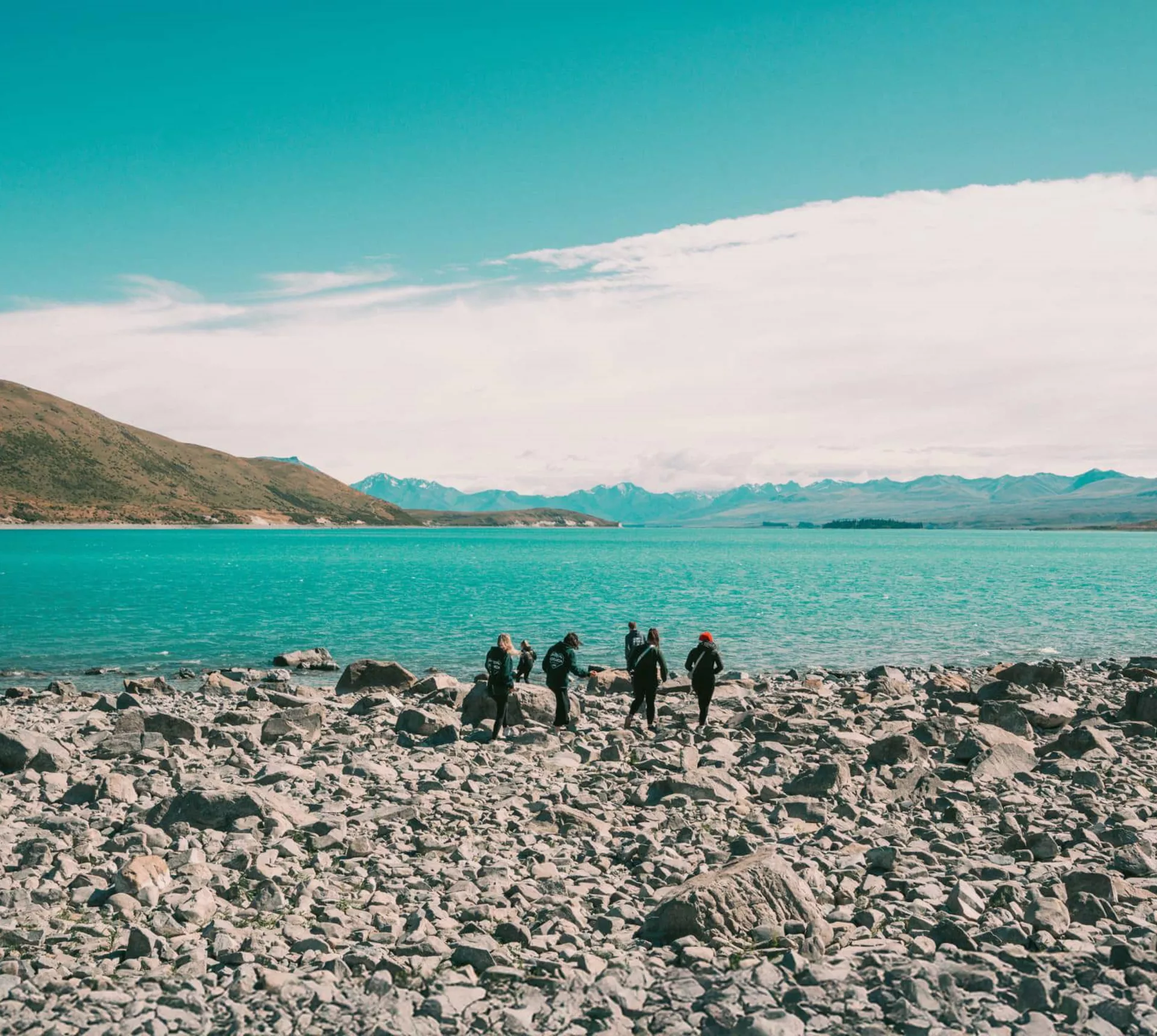 Group of travellers walking along the rocky shoreline of glacial Lake Pukaki with the vivid turquoise water and snow-capped Southern Alps in the background New Zealand - Contiki Best of NZ Tour