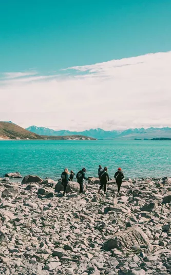 Group of travellers walking along the rocky shoreline of glacial Lake Pukaki with the vivid turquoise water and snow-capped Southern Alps in the background New Zealand - Contiki Best of NZ Tour