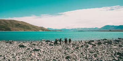 Group of travellers walking along the rocky shoreline of glacial Lake Pukaki with the vivid turquoise water and snow-capped Southern Alps in the background New Zealand - Contiki Best of NZ Tour