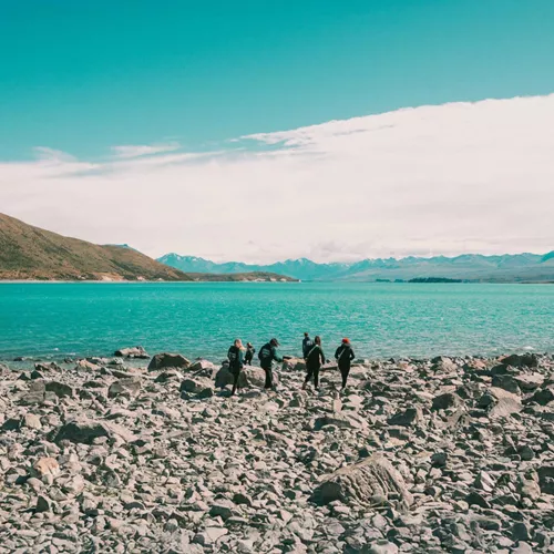 Group of travellers walking along the rocky shoreline of glacial Lake Pukaki with the vivid turquoise water and snow-capped Southern Alps in the background New Zealand - Contiki Best of NZ Tour