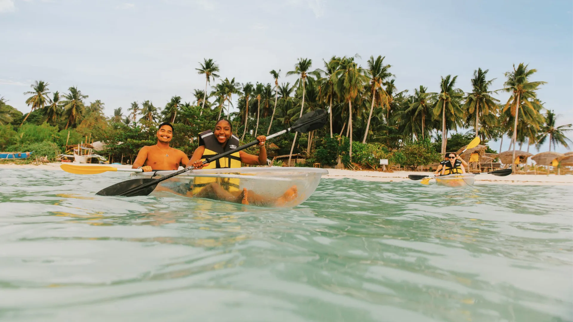 Two young travellers kayaking in a transparent kayak in crystal clear waters off a palm-lined beach in Palawan Philippines - Philippines Island Hopping West Tour