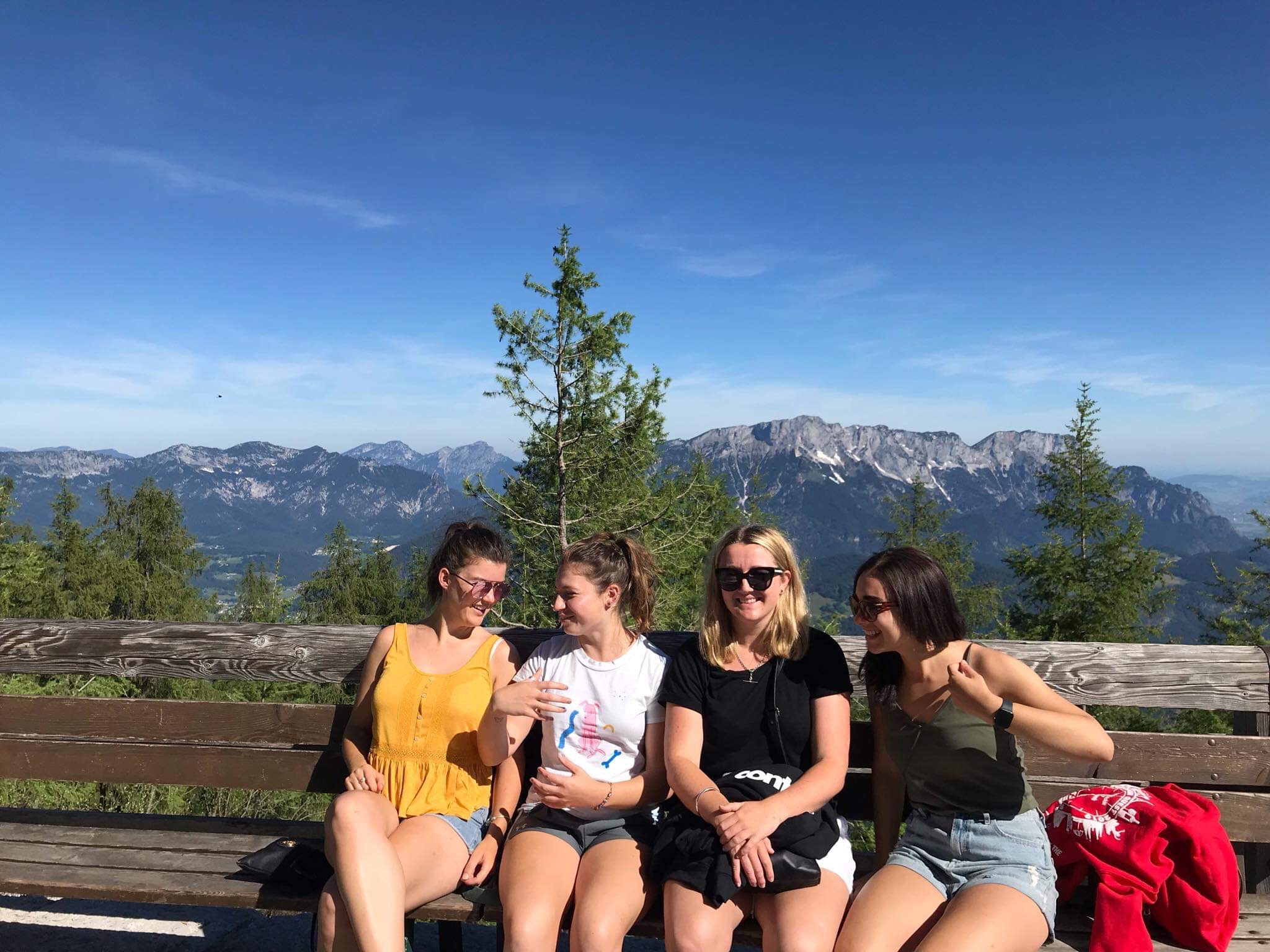 Four young friends sitting on a bench with a forest and mountains in the background.