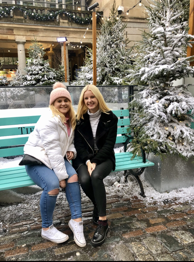 BFFs Imogen and Jess sitting on a bench surrounded by pine trees covered in snow.
