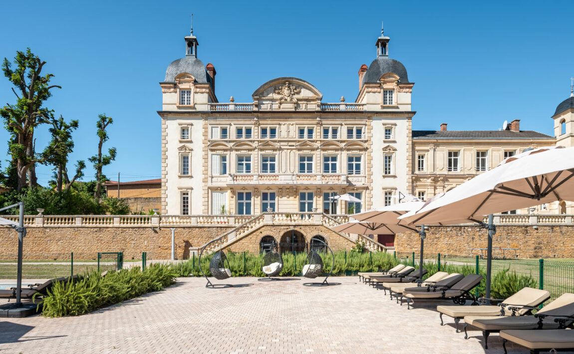 A large stone Contiki Château building is in the middle of a courtyard with lounge chairs and umbrellas.