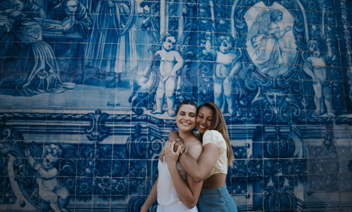 Two women hugging during their travel adventure in front of a blue tiled wall.