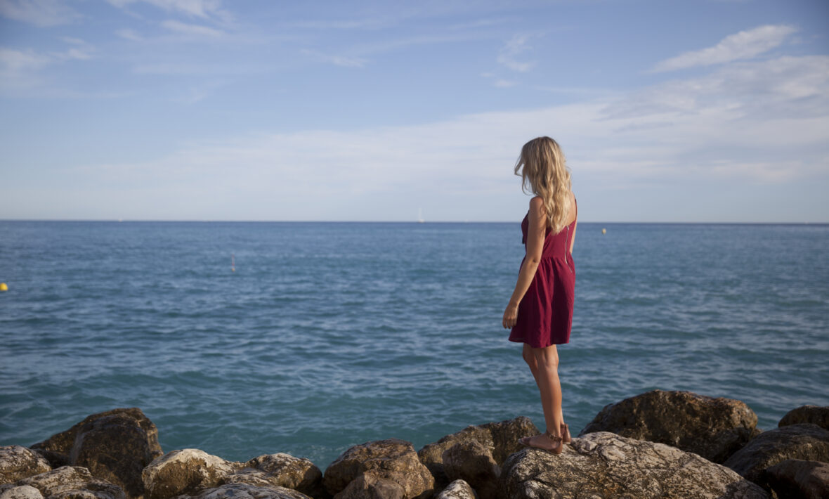 A woman in a red dress standing on rocks near the ocean, portraying depression.