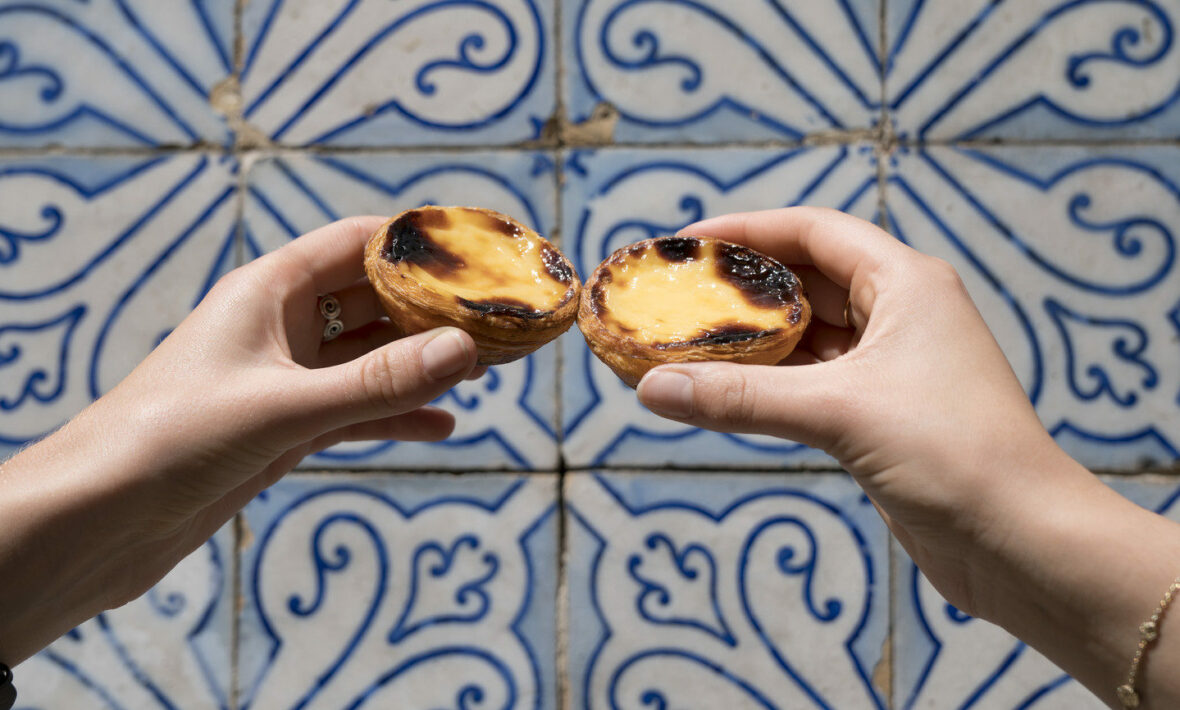 A pair of hands holding two pastries in front of a blue and white tiled wall in Portugal.