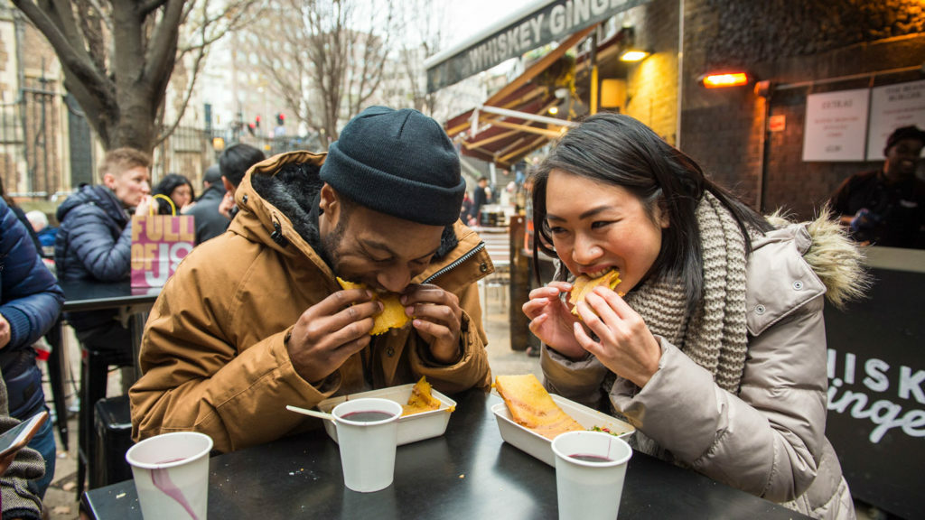 Two people enjoying food at an outdoor table at a food festival.