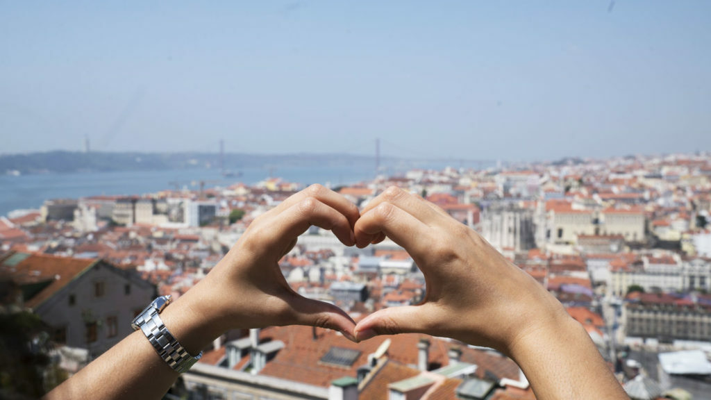 A person's hands form a heart shape over the city of Lisbon, emphasizing self-love.