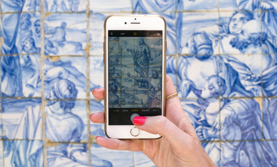 A person using travel apps on a cell phone in front of a blue tiled wall.