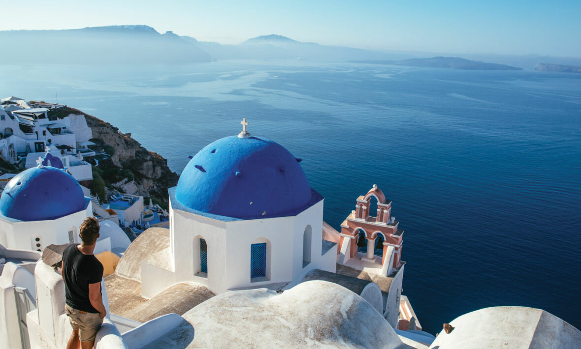 A man with a travel style is standing on a cliff overlooking the ocean.