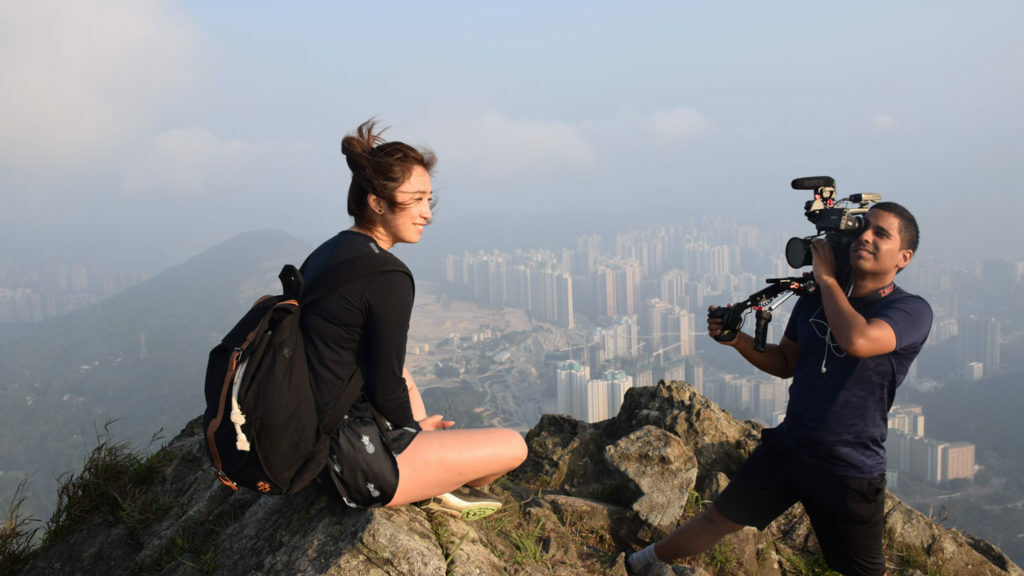 A woman, a travel filmmaker, sitting on top of a mountain with a camera.