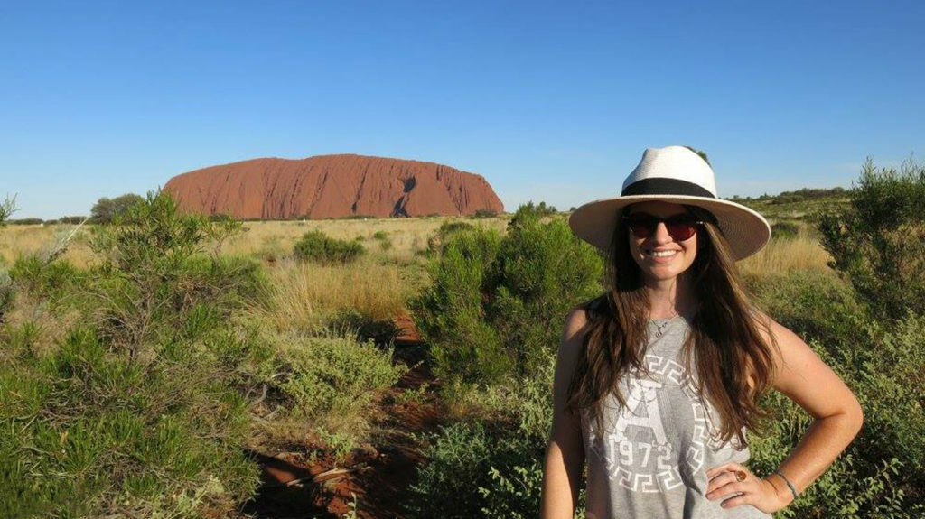 A woman in a hat traveling in front of Uluru.