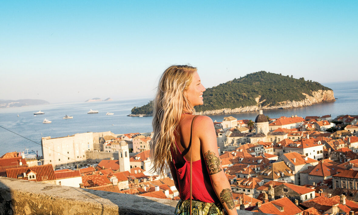 A woman quit the 9-5 and is standing on top of a wall overlooking the city of Dalmatia.