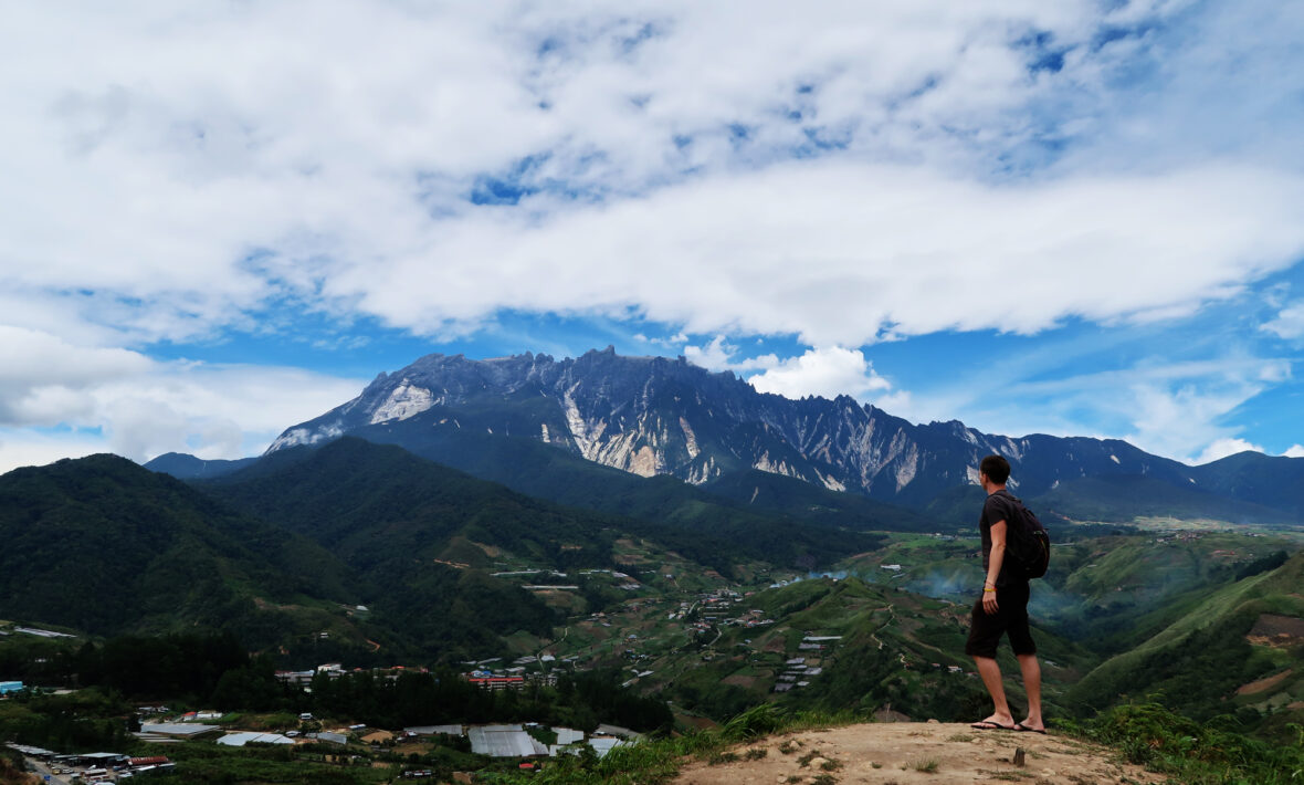 A man backpacking on a budget standing on top of a mountain overlooking a village.