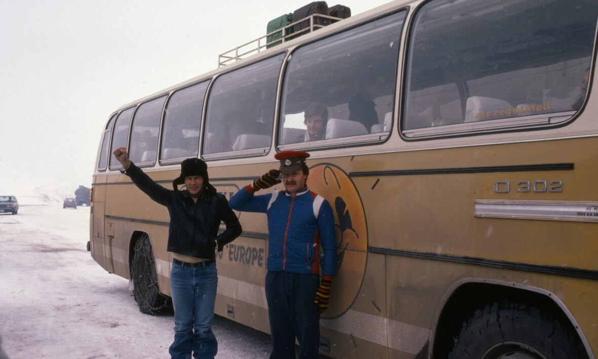 Two people standing in front of a retro yellow bus, reminiscent of a contiki ad.