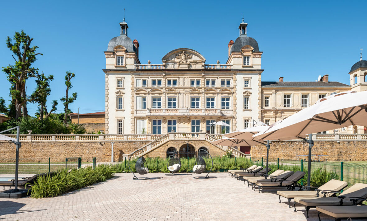 A large stone Contiki Château building is in the middle of a courtyard with lounge chairs and umbrellas.