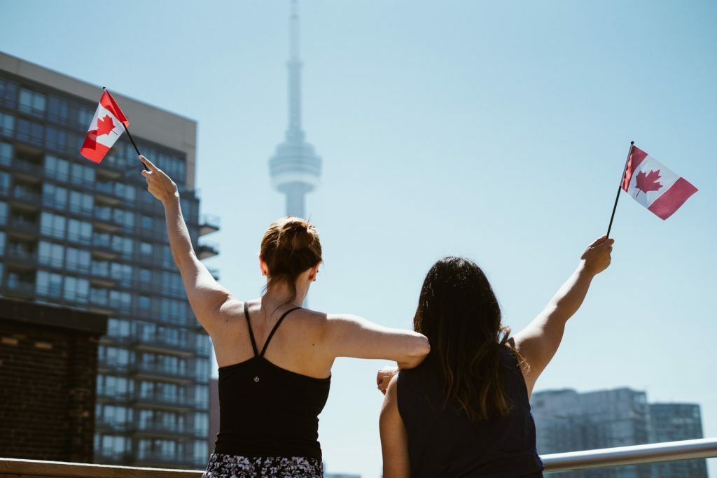 Two women waving Canadian flags in front of the CN Tower in Toronto encounter offensive remarks about Canada.