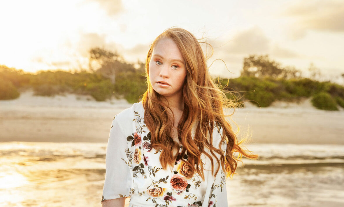 A girl in a floral dress standing on the beach at sunset, Madeline Stuart.