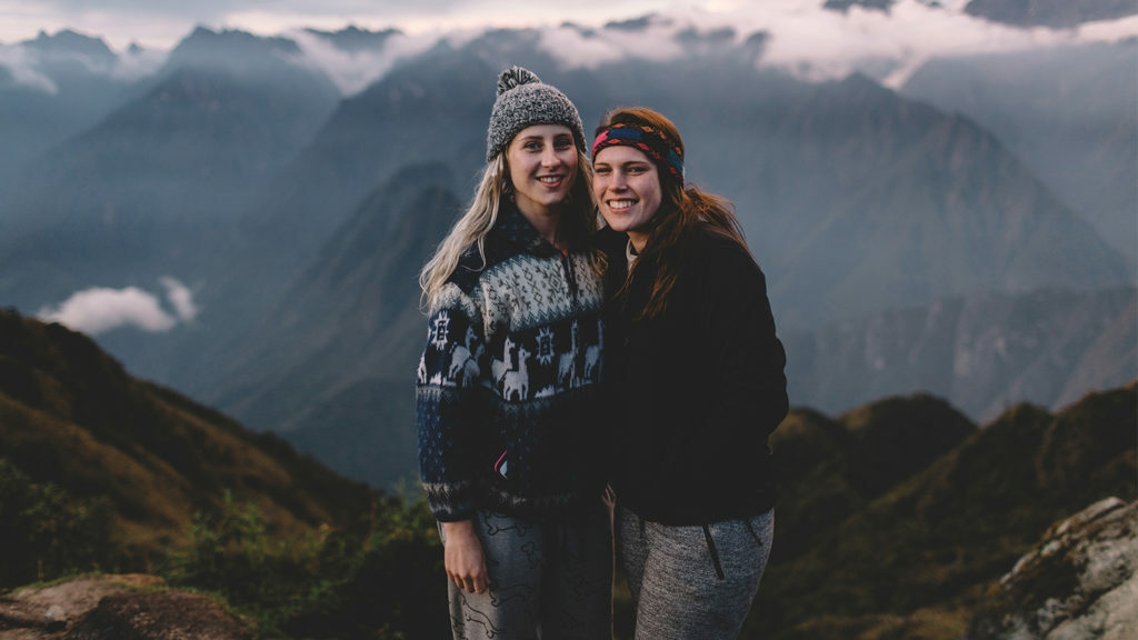 Two millennial women standing on top of a mountain with mountains in the background.