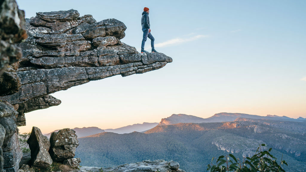 A man with a side hustle standing on top of a cliff in the blue mountains.