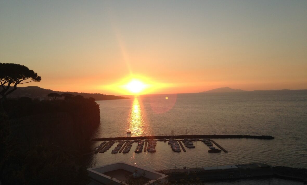 Travel to Italy and watch the sun setting over a charming harbor with boats docked.