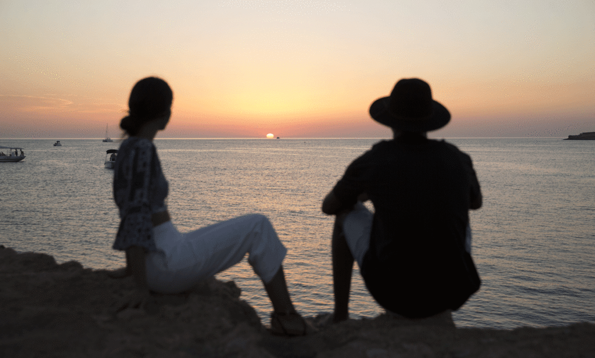 Two people experiencing true love while sitting on a cliff overlooking the ocean at sunset.